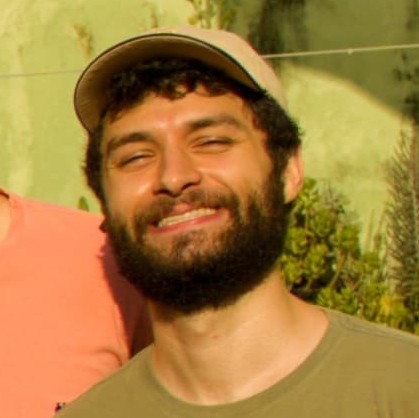 A picture of me. I am smiling, with a beard and curly hair, wearing a light National Geographic cap and a green shirt, standing outdoors against a greenish-yellow background with soft sunlight.