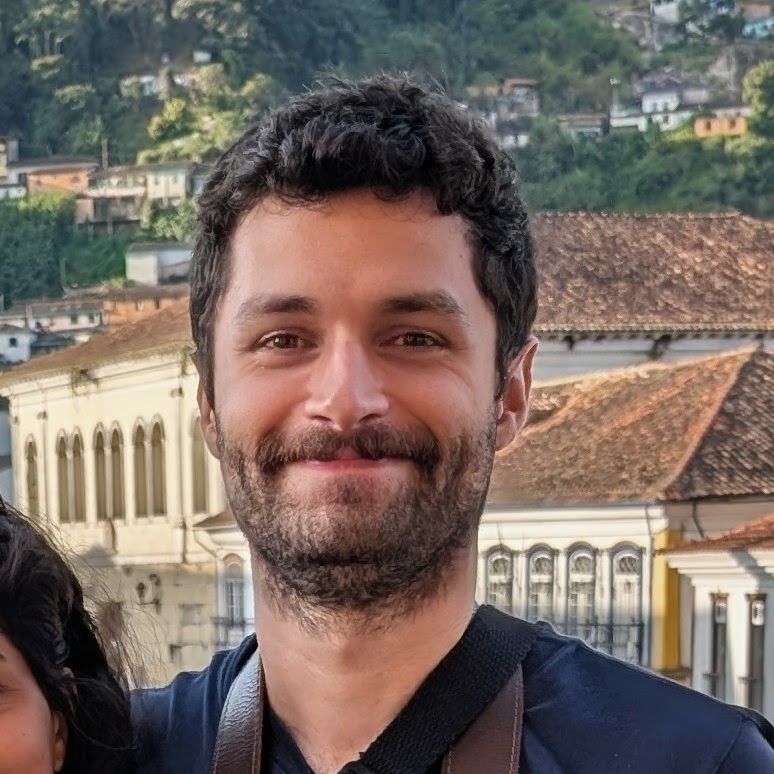 A picture of me. I am smiling, with a beard and curly hair, wearing a dark shirt. Behind me there are colonial-style buildings with white walls and terracotta roofs, and green hills in the background.